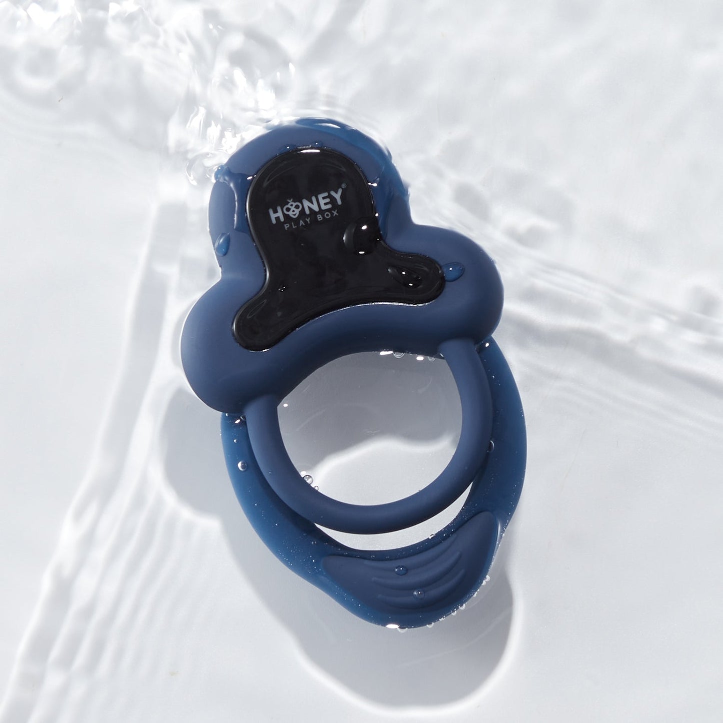 A dark blue Anello Honey Play Box cock ring with water droplets on it, laying on a white background
